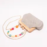 Cartera de mano tipo clutch con estructura de madera clara y cuerpo de tela tejida en tonos beige y plateado. Posee una correa larga de cadena dorada con cuentas facetadas de colores variados (naranja, blanco, rosa, turquesa, amarillo, verde). Incluye dos esferas de madera como detalle decorativo en el marco de madera.