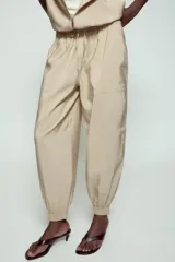 Pantalón satinado de tiro alto color beige, con cintura elástica ajustable con cordones, bolsillos delanteros y puños elásticos en los tobillos.