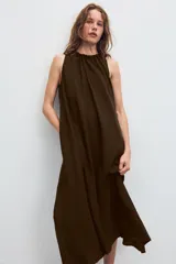 Vestido midi de color marrón con acabado satinado, cuello redondo con detalle fruncido y diseño sin mangas. Presenta un corte fluido con bajo asimétrico y bolsillos laterales ocultos.