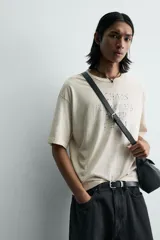 Camiseta de corte holgado y estilo cropped, color beige con efecto lavado. Presenta cuello redondo, mangas cortas y una estampa frontal con texto en azul que simula trazos de rotulador.