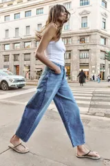 Jeans de tiro alto con diseño barrel, confeccionados en denim azul. Presentan cinco bolsillos, cierre frontal con botones y detalle de costuras verticales en el frente y la espalda. Su corte es holgado en la pernera y se estrecha hacia el tobillo.