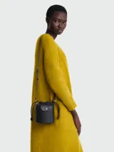 Bolso pequeño tipo cubo (bucket bag) de cuero negro, con cordón ajustable en la parte superior y solapa con broche a presión. Se lleva cruzado con una correa fina y ajustable.