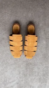 Zuecos de punta cuadrada, confeccionados en gamuza beige con diseño de tiras entrelazadas tipo huarache y suela plana marrón.