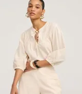 Blusa estilo bata color blanco roto, confeccionada en cambraia de algodón y lino. Presenta cuello con lazo ajustable, canesú fruncido en el busto y mangas tres cuartos con puños elásticos y bordados.