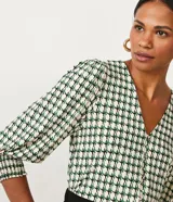 Blusa de jersey con estampado geométrico en tonos blanco, negro y verde. Tiene escote en V, mangas 3/4 abullonadas con elástico en los puños y cierre frontal con botones.