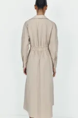 Vestido midi de color beige con cuello solapa y escote en pico. Presenta mangas largas, cintura elástica en la espalda y un cinturón a contraste con hebilla metálica. Incluye bolsillos laterales ocultos.
