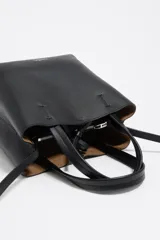 Bolso pequeño de piel vacuna color negro, con estructura rígida, doble asa de mano y correa larga ajustable para llevar al hombro o cruzado. Presenta cierre de botón a presión y logo metálico de la marca en el frente.