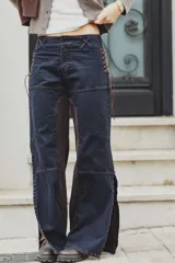 Jean de corte recto en denim azul oscuro con paneles laterales en contraste color marrón. Presenta detalles de cordones trenzados en los laterales de la cintura, costuras horizontales en las piernas y tajos en el ruedo.