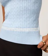 Top sin mangas de punto color celeste claro, con cuello polo ribeteado con dos líneas blancas y cierre frontal de tres botones. El tejido presenta un patrón de trenzas verticales.