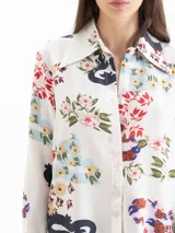 Camisa de seda color blanco con estampado floral y de dragones en tonos rojos, azules y verdes. Tiene cuello clásico, mangas largas con puños ajustables y botones forrados.