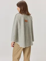 Capa de tejido de punto en color gris melange, con diseño abierto y cierre frontal mediante un botón tipo duffle de madera con detalle en cuero. Presenta bolsillos laterales amplios y bordes con costuras a la vista.