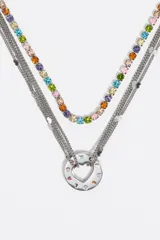 Collar de estilo gargantilla y largo, compuesto por múltiples cadenas plateadas. Dos de las cadenas están adornadas con cristales de colores brillantes (rosa, naranja, amarillo, verde, azul, violeta). Las otras cadenas son de eslabones finos. El dije principal es un aro metálico plateado con un corazón calado en el centro, decorado con pequeños cristales de colores.