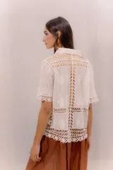 Camisa de algodón color crudo, con cuello camisero, mangas cortas y cierre frontal con botones. Presenta paneles de tejido calado y detalles bordados transparentes.