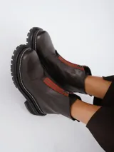 Botinetas de cuero color marrón oscuro con suela track negra y detalle de cierre frontal con cinta en color naranja.