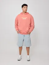 Canguro con capucha color salmón/coral desgastado, con efecto lavado. Presenta un estampado frontal con el texto "RUSTY & SUN" y un diseño de olas en color amarillo pálido. Tiene bolsillo canguro frontal y puños y cintura acanalados.