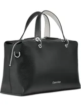 Cartera tipo satchel de Calvin Klein en color negro, confeccionada en material sintético con textura granulada. Presenta doble asa de mano, correa de hombro ajustable y extraíble, y cierre superior con cremallera. Incluye accesorios colgantes decorativos: un monedero circular y un pequeño estuche con solapa, ambos con el monograma de la marca.