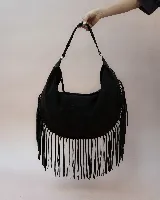 Cartera tipo hobo de gran tamaño, confeccionada en gamuza negra, con diseño de flecos largos en los laterales y la base. Presenta un asa trenzada con detalles artesanales y cierre superior.