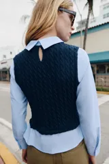 Camisa de popelín celeste con cuello solapa y manga larga, combinada con un chaleco de punto trenzado en color azul marino superpuesto. Presenta un diseño de capas con cierre en la espalda mediante botones ocultos.