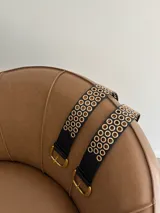 Cinturón negro de cuero ecológico con hebilla metálica dorada y doble hilera de ojales dorados a lo largo de toda la tira.