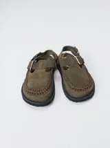 Slipper tipo guillermina confeccionada en gamuza color verde oliva, con plataforma de goma negra y doble hebilla ajustable. Presenta un pespunte decorativo en contraste en el borde inferior.