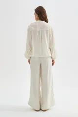 Blusa de algodón color beige, con cuello redondo y cierre frontal con botones. Presenta delicados bordados decorativos en tono tostado sobre el pecho y los hombros, mangas largas abullonadas con puños ajustados y diseño holgado.