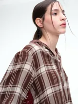 Camisa de corte relajado con estampado de cuadros en tonos negro y beige. Presenta cuello camisero, cierre frontal parcial con botones y mangas largas con puños ajustables mediante lazos. El diseño destaca por un detalle de encaje en la espalda que aporta contraste.