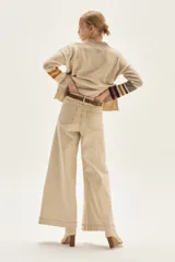 Pantalón de jean color beige claro, tiro alto, de corte ancho (wide leg) y largo hasta el tobillo con dobladillo. Presenta costuras verticales marcadas en el frente de las piernas.