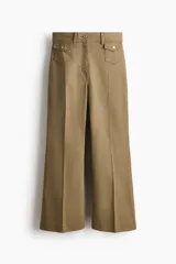 Pantalón de sarga color tostado, de tiro alto y corte acampanado con diseño tobillero. Presenta bolsillos frontales tipo parche con solapa abotonada y pliegues marcados al frente y en la espalda.
