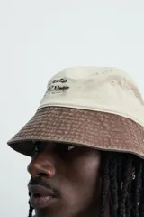 Sombrero bucket confeccionado en tejido de algodón con efecto lavado, diseño bicolor en tonos beige claro y marrón oscuro. Presenta ala ancha con pespuntes y un bordado frontal en contraste.