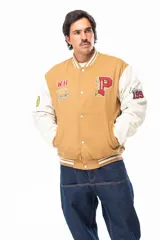 Campera estilo varsity con cuerpo color camel y mangas blancas. Presenta cierre frontal con botones a presión, cuello, puños y ruedo con franjas elásticas a rayas. Incluye parches bordados en el pecho y las mangas con motivos universitarios.
