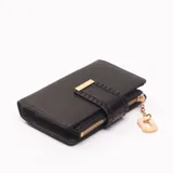 Billetera negra de textura granulada con cierre perimetral y solapa frontal decorativa con detalle metálico dorado. Incluye un dije colgante con forma geométrica en tono crema.