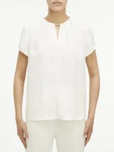 Blusa blanca de poliéster reciclado con cuello redondo, mangas cortas y cierre con cremallera en la espalda. Presenta una abertura en la parte delantera con barra metálica.