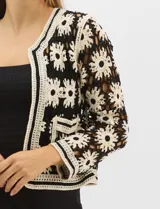 Cardigan abierto de tejido crochet con diseño de flores bicolores en negro y crema. Presenta mangas largas, frente abierto sin cierre y bolsillos frontales tipo parche. Posee terminaciones con franjas a contraste en escote, puños y ruedo.