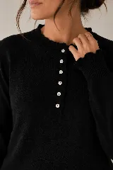 Sweater de punto negro en doble hilado Bremer, con cuello redondo y cierre frontal parcial de botones blancos con ribetes ondulados, mangas largas con puños elastizados y volados sutiles en el cuello y los puños.