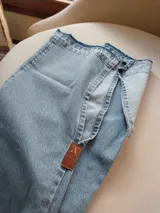 Pantalón de jean celeste con cintura elastizada, corte recto, bolsillos laterales y aplique de cuero marrón en el frente. Presenta botones metálicos en los laterales y etiqueta de cuero en la parte posterior.