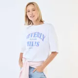 Camiseta de corte oversize color blanco, con cuello redondo y mangas cortas. Presenta un estampado frontal con la inscripción 'Beverly Hills L.A. 1989 West Co' en tonos azules.