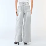 Jean de corte wide leg y tiro alto, confeccionado en denim de color gris lavado. Presenta pinzas en la cintura, cierre frontal con botón y bolsillos laterales.