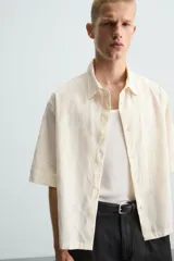 Camisa de corte relajado (relaxed fit) color crema o blanco roto, confeccionada en tejido con mezcla de algodón. Presenta un bordado sutil en relieve con patrón de ondas o líneas diagonales entrecruzadas. Tiene cuello solapa, manga corta y un bolsillo de plastrón en el pecho.