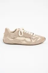 Zapatilla de cuero legítimo en tonos beige y crema, con diseño de flecos en el lateral y costuras decorativas visibles en el contorno. Presenta cordones al tono y suela plana.