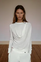 Jersey de punto fluido color blanco, con cuello redondo, manga larga y detalle de pliegues drapeados en el hombro.