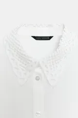 Camisa blanca de manga larga con cuello de solapa y detalles de encaje a tono en el cuello y los puños. Presenta cierre frontal con botones y corte recto.