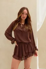 Vestido corto marrón de tejido semitransparente, con mangas largas abullonadas, escote redondo con cordones para ajustar y falda con volados en capas.