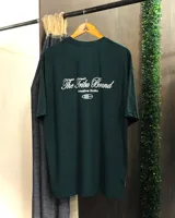 Remera de algodón color verde oscuro, corte oversized, con estampado en la espalda que dice "The Tribu Brand Creative Studio" en tipografía cursiva blanca, acompañado de un pequeño logo.