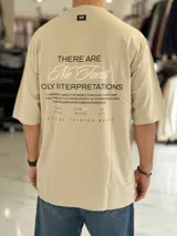 Remera oversize color beige, con mangas cortas y cuello redondo. Presenta un estampado tipográfico en la espalda con la frase "There are no facts, only interpretations" y otros detalles de diseño.