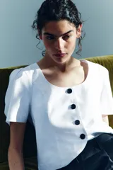 Blusa corta blanca con escote en U, mangas cortas abullonadas y cierre frontal con botones negros contrastantes.