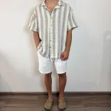 Camisa de manga corta con corte oversize, cuello tipo cubano y cierre frontal con botones. Presenta un diseño de rayas verticales en tonos crudo y gris.