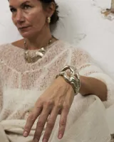 Brazalete abierto de diseño orgánico y voluminoso, con acabado metálico brillante. Su forma irregular y ergonómica permite un ajuste cómodo en la muñeca o antebrazo.