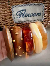 Set de cinco brazaletes de diferentes materiales y colores: tres brazaletes rígidos de resina en tonos naranja y beige marmolado, un brazalete de madera marrón con apliques de estrellas y círculos dorados, y un brazalete de madera marrón liso.