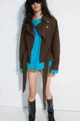 Cazadora trench corta color marrón, confeccionada en mezcla de algodón, con cuello solapa, cierre cruzado con botones, bolsillos de vivo en el delantero y cinturón ajustable del mismo tejido.