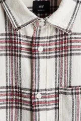 Camisa de franela de algodón suave con efecto lavado, de corte relajado y cuello inglés. Presenta un estampado a cuadros en blanco, rojo y negro, tapeta clásica de botones, un bolsillo superior abierto y canesú en la espalda. Tiene mangas largas con tapeta, puños abotonados y bajo levemente redondeado.
